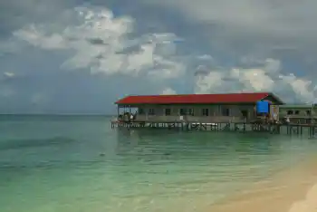 Votre hébergement simple et économique au-dessus de l'eau. La longhouse du Mabul Backpackers perchée au-dessus d'une mer calme, vue depuis la plage de sable sous un ciel nuageux spectaculaire.