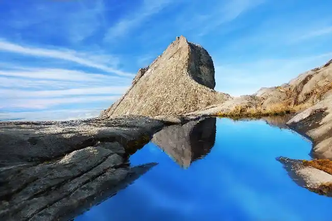 Stunning reflection of St. John's Peak in a still pool of water on the summit of Mount Kinabalu under a clear blue sky.