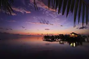 Coucher de soleil aux teintes violettes et orangées sur un resort sur pilotis de la région de Sipadan, avec les lumières de la jetée se reflétant sur la mer.