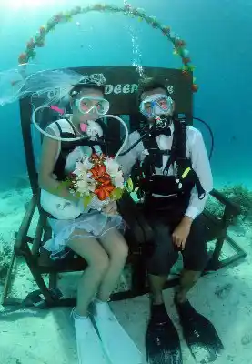 Eine wirklich einzigartige Art, Ihren Hochzeitstag zu feiern. Abenteuerlustiges Paar in voller Tauchausrüstung feiert seine einzigartige Unterwasser-Hochzeit, komplett mit Brautstrauß und einem Blumenbogen auf dem Meeresgrund.