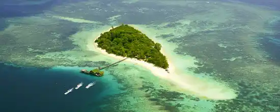 See the island's beauty from a bird's-eye view. Breathtaking sky view of Lankayan Island, showing the lush island, extensive coral reef, and dive boats heading out for an adventure in the Sulu Sea.