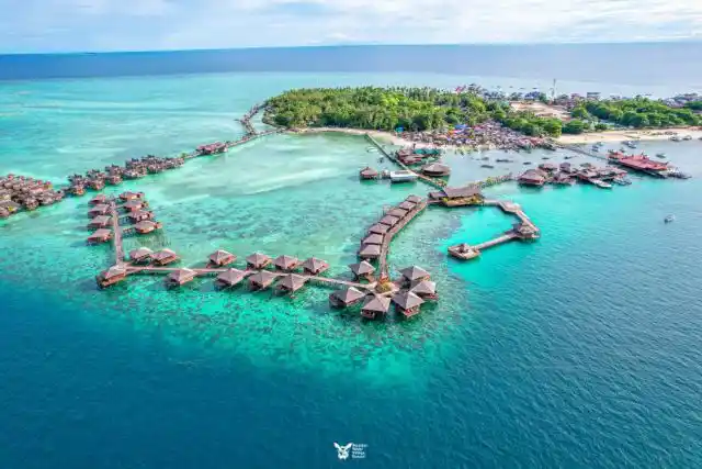 See the entire island and its resorts from above. Spectacular high-altitude drone panorama of Mabul Island, showing the luxurious Sipadan Water Village in the foreground and the rest of the island's resorts and villages.